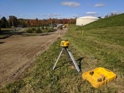 surevey machine on tri-pod aimed down the dirt road, while on grassy knol, 
                water tank in far background