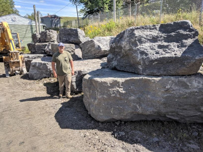 man standing to left of limestone blocks. Two blocks are taller then man.
                Excavator in background