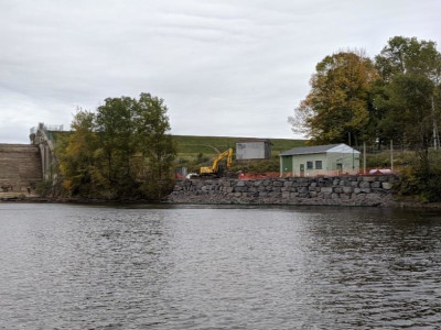 view of completed wall across the water, excavator and building in far back above wall