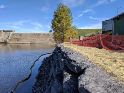view of completed wall to right, orange security fence, water to the left, tree and dam in background