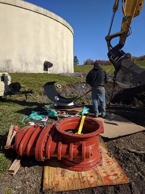 tank in background, man and excavation claw in center, 
                large red pipe-sleeve on plywood in foreground