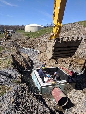 2 men in cement pit with pipe through center being lowered by excavation claw